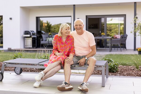 Ein Paar sitzt entspannt auf einer Gartenliege vor einem modernen Haus und lächelt einander an.