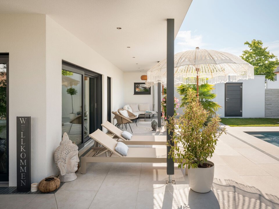 Überdachte Terrasse mit Liegen, Sonnenschirm und Blick in den Garten.