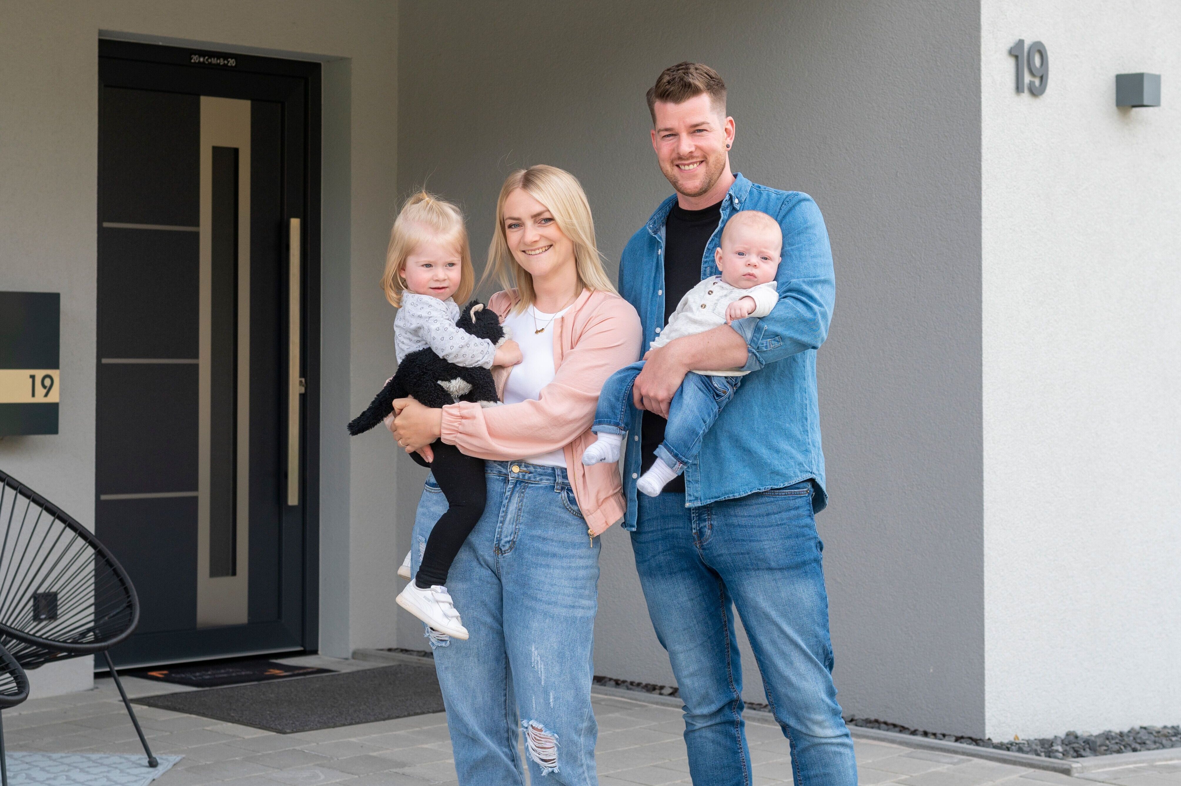 Familie mit zwei kleinen Kindern steht vor modernem Fertighaus mit Hausnummer 19 und dunkler Haustür