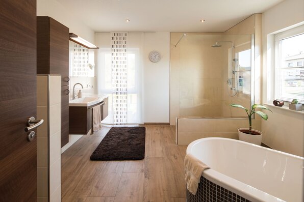Modernes Badezimmer in einem frei geplanten FingerHaus mit freistehender Badewanne, bodentiefer Glasdusche, Doppelwaschbecken, Holzoptik-Boden und großen Fenstern.