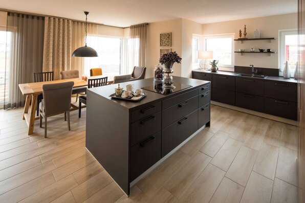 Essbereich und Küche im FingerHaus MEDLEY 300 B Musterhaus Berlin/Werder mit dunkler Kücheninsel, Esstisch aus Holz und großen Fenstern.