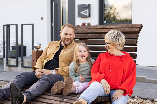 Drei Generationen sitzen gemeinsam auf einer Holzbank vor dem Kundenhaus NIVO 80 und lachen miteinander.