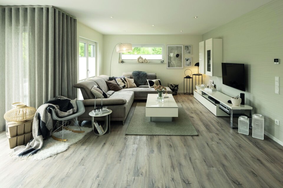 Wohnzimmer mit großer Eckcouch, Couchtisch und Fernseherwand. Helle Wände, Vorhänge und Holzfußboden sorgen für eine freundliche Atmosphäre.