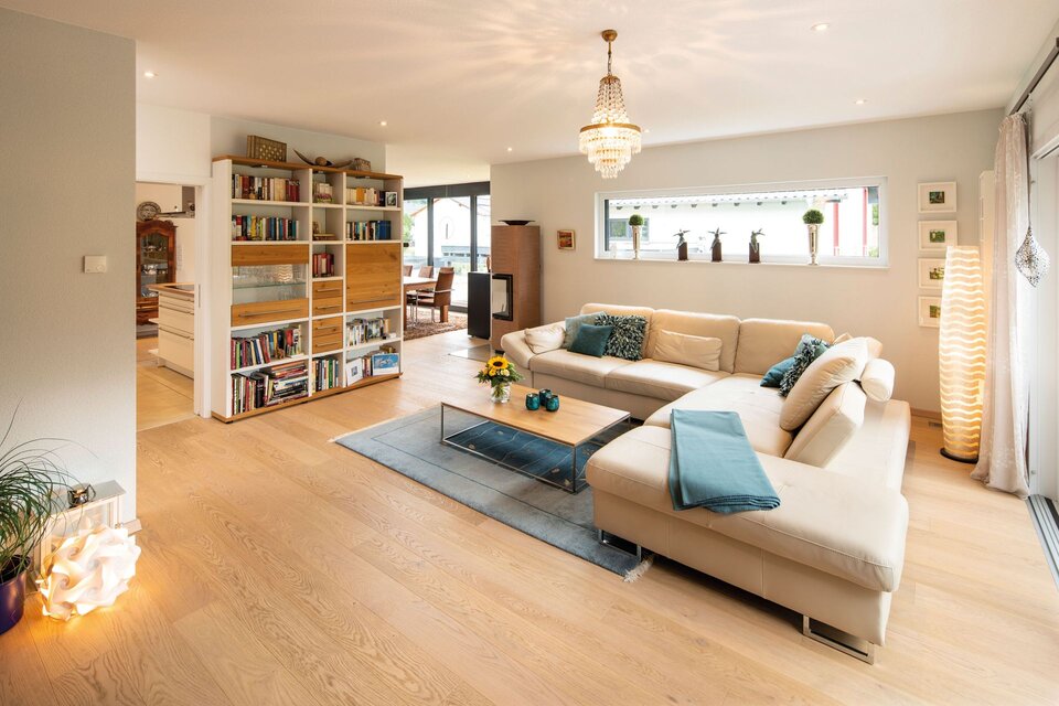 Gemütliches Wohnzimmer in einem frei geplanten FingerHaus mit heller Eckcouch, Bücherregal, Glastisch, Holzboden und großen Fenstern.
