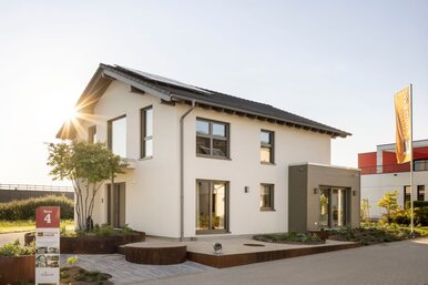 Außenansicht des FingerHaus Musterhauses ARTIS 300 in Nürnberg mit weißer Putzfassade, dunkel gedecktem Satteldach und seitlichem Anbau mit horizontaler Fassadenverkleidung; davor gepflasterter Weg, Hochbeete mit Bepflanzung, Bäume, Fahnenmasten und tief stehende Sonne.