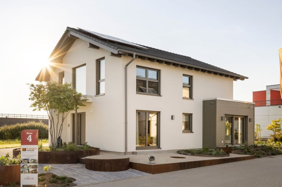 Außenansicht des FingerHaus Musterhauses ARTIS 300 in Nürnberg mit weißer Putzfassade, dunkel gedecktem Satteldach und seitlichem Anbau mit horizontaler Fassadenverkleidung; davor gepflasterter Weg, Hochbeete mit Bepflanzung, Bäume, Fahnenmasten und tief stehende Sonne.