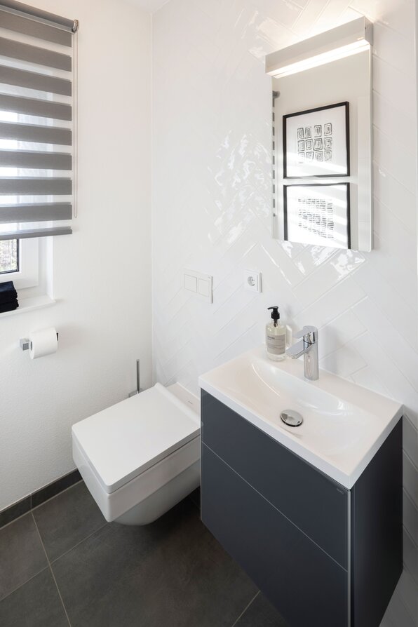 Modernes Gäste-WC im Musterhaus TALO Fulda mit dunklem Waschtisch, weißem Waschbecken und wandhängendem WC, gestaltet in klaren Linien und zeitlosem Design.