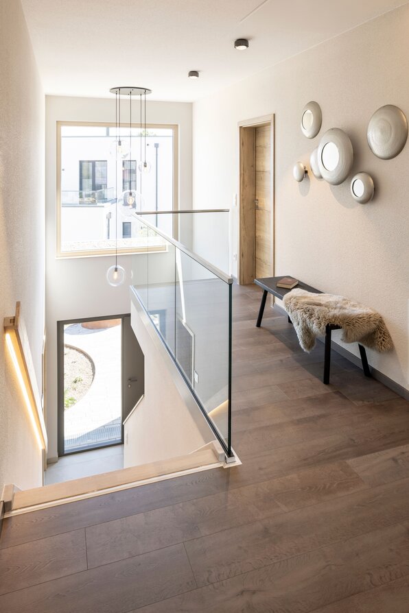 Heller Flur im Obergeschoss des FingerHaus ARTIS 300 Musterhauses Nürnberg mit Glasgeländer, Holzfußboden und moderner Beleuchtung.