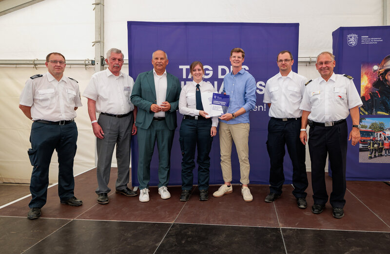 Sieben Personen stehen bei einer Preisverleihung in einem Veranstaltungszelt vor einem violetten Hintergrund. Zwei Personen in der Mitte halten gemeinsam eine Urkunde mit der Aufschrift „Arbeitgeber-Award 2026“, mehrere Personen tragen Uniform.