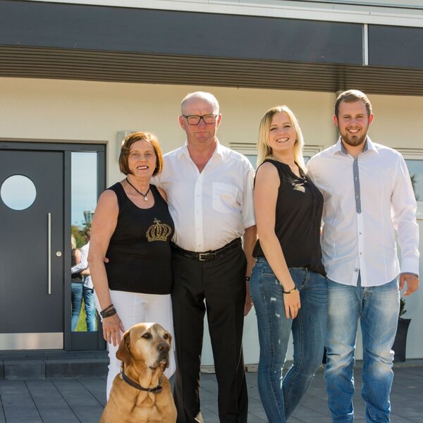 Vierköpfige Familie mit Hund steht vor dem Eingang ihres modernen FingerHaus mit dunkler Haustür und heller Fassade.