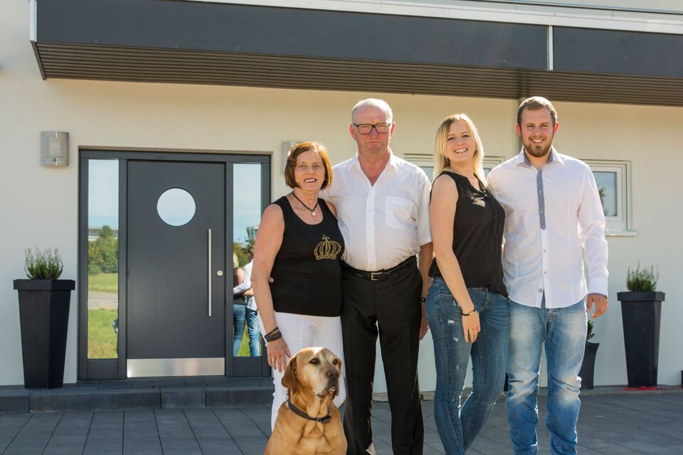 Vierköpfige Familie mit Hund steht vor dem Eingang ihres modernen FingerHaus mit dunkler Haustür und heller Fassade.