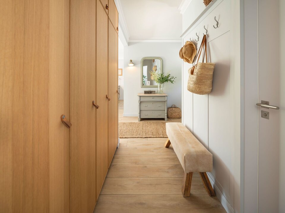 Flur mit großer Einbauschrankfront aus Holz, Bank, Wandhaken und Spiegelkommode im Hintergrund.
