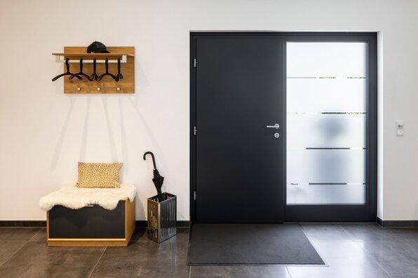 Moderner Eingangsflur in einem frei geplanten FingerHaus mit schwarzer Haustür, seitlichem Glaselement, Sitzbank mit Kissen und Wandgarderobe. Ein Schirmständer ergänzt den funktionalen Bereich.