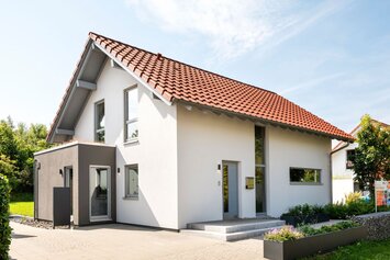 Außenansicht eines frei geplanten Finger-Haus Musterhauses in Marburg mit weißer Putzfassade, rotem Satteldach und dunkelgrauem Anbau; davor gepflasterte Terrasse, Hochbeete mit Bepflanzung, Rasenfläche und eine FingerHaus-Fahne am Grundstück.