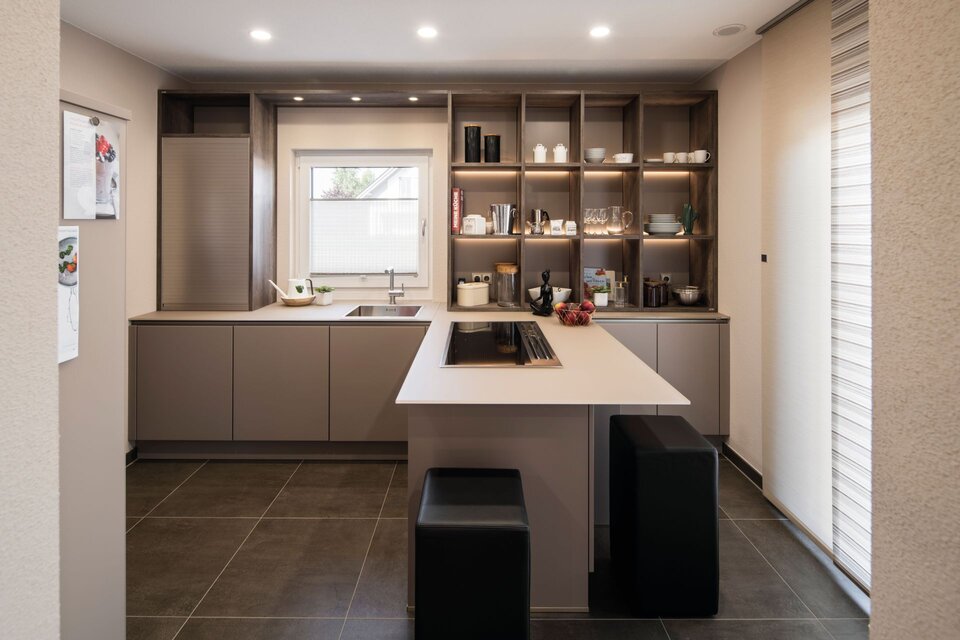 Moderne Küche mit großer Kochinsel, grauen Fronten und dunklem Fliesenboden. Offenes Holzregal mit Beleuchtung an der Rückwand, zwei schwarze Hocker an der Insel und Fenster mit Lamellenrollo im Musterhaus UNO Leipzig.