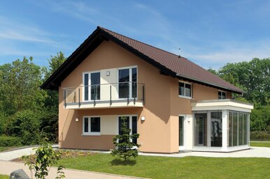 Zweistöckiges Fertighaus mit Balkon, Wintergarten und braunem Satteldach auf gepflegtem Rasen mit jungen Bäumen