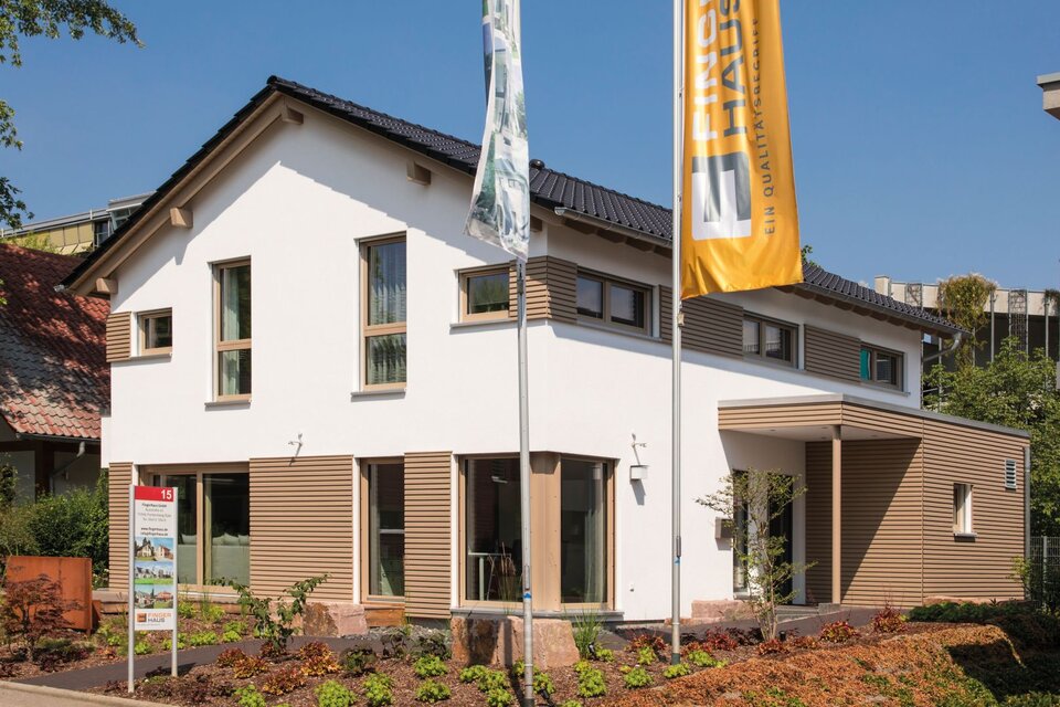 Außenansicht des Musterhauses MEDLEY in Fellbach mit heller Putzfassade, Holzdetails und großen Fenstern, ergänzt durch Flaggenmasten mit FingerHaus-Beschriftung.