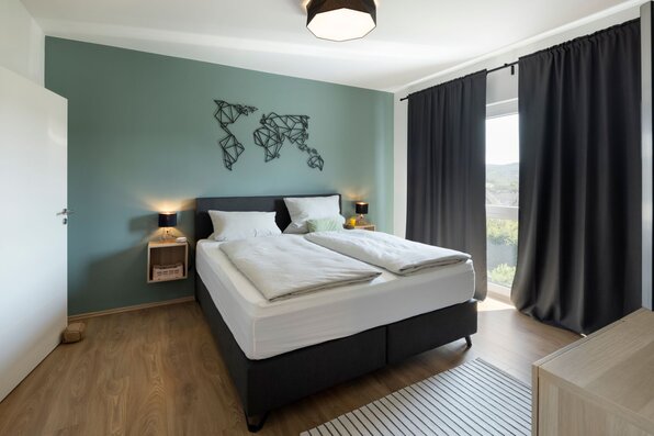 Schlafzimmer mit Doppelbett, dunkelgrüner Akzentwand und großem Fenster mit Vorhängen. Holzfußboden, Nachttische mit Lampen und dezente Dekoration.