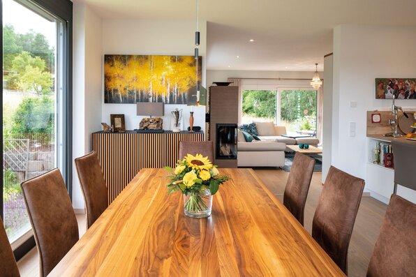 Heller Essbereich in einem frei geplanten FingerHaus mit großem Holztisch, braunen Stühlen, modernen Bildern und Blick ins Wohnzimmer mit Kamin und Sofaecke.
