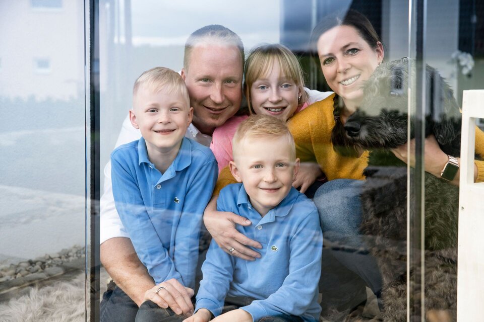 Familie sitzt mit ihrem Hund hinter einer großen Glasscheibe und blickt lächelnd nach draußen.