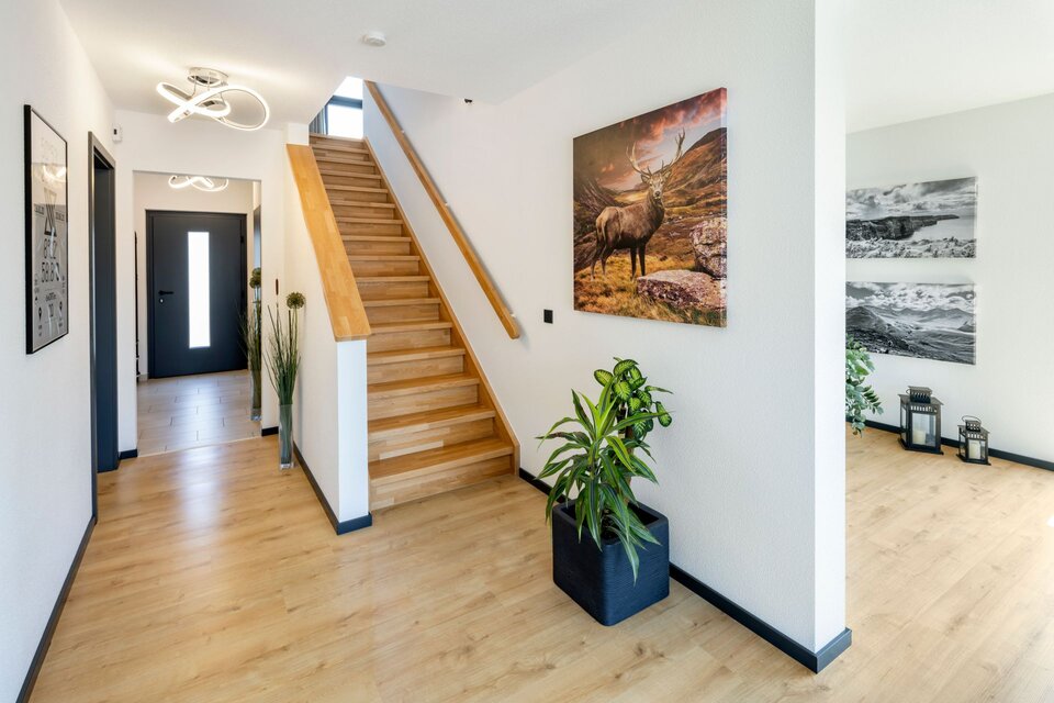 Heller Eingangsbereich mit geschlossener gerader Holztreppe, weißen Wänden und Pflanzen. Moderne Deckenleuchte, helle Fliesen und offene Raumgestaltung.