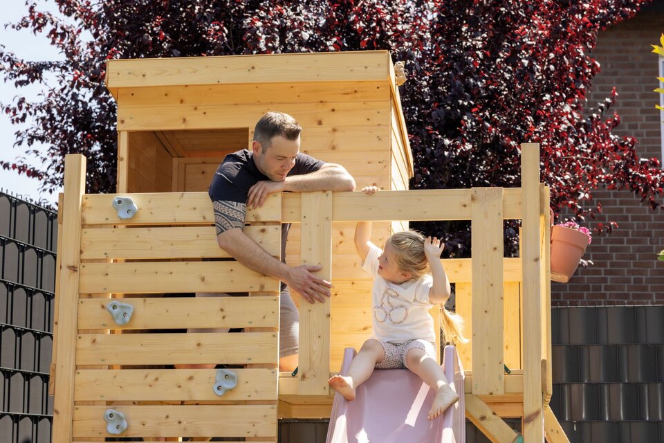 Vater und Kind spielen auf einem Holzspielhaus mit Rutsche im Garten