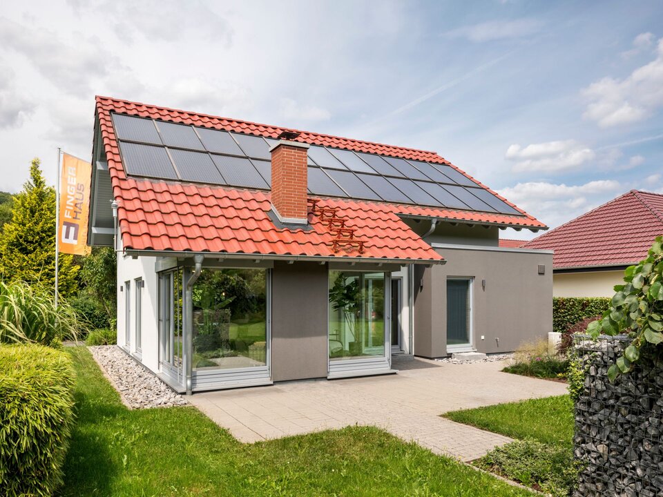 Außenansicht des frei geplanten FingerHaus Musterhauses in Marburg mit rotem Satteldach, Photovoltaikanlage und großem Wintergarten.