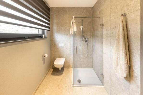 Modernes Bad im FingerHaus ARTIS 300 Musterhaus Würzburg mit bodengleicher Dusche, heller Natursteinoptik und WC.