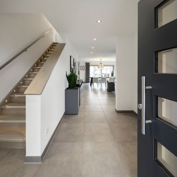 Moderner Eingangsbereich im Musterhaus NEO 312 in Bad Vilbel mit gefliesten Böden, stilvoll beleuchteter Treppe und Blick ins Esszimmer.