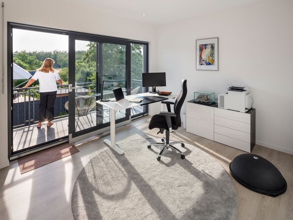 Arbeitszimmer mit Schreibtisch, Bürostuhl und einer Person auf dem Balkon vor bodentiefen Fenstern.