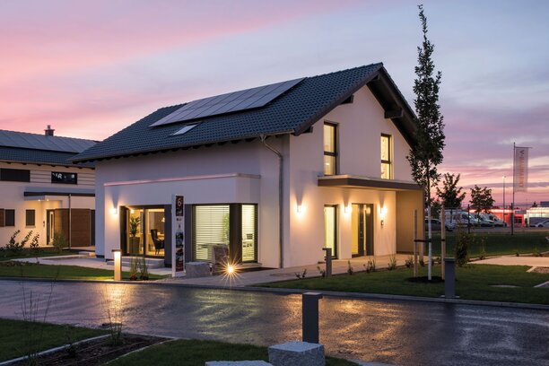 Abendliche Außenansicht des FingerHaus-Musterhauses NEO in Günzburg mit beleuchteten Fenstern, moderner Architektur und Photovoltaikanlage auf dem Satteldach.