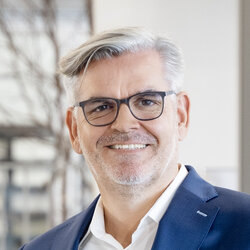 Mann Michael Müller mit grauen Haaren trägt einen dunkelblauen Anzug und weißes Hemd vor modernem Innenraum mit Holz- und Glasakzenten