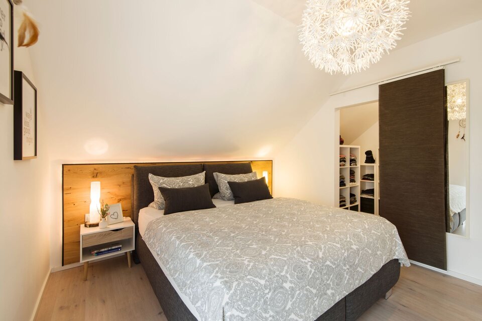 Gemütliches Schlafzimmer mit Doppelbett, Holzrückwand und Nachttischen mit Lampen. Dachschräge, heller Boden und dekorative Leuchte an der Decke.