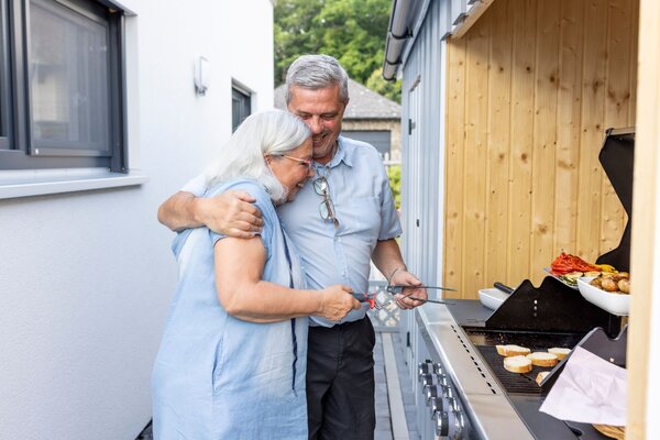 Älteres Paar grillt gemeinsam an einem Gasgrill neben einem modernen Flachdachhaus