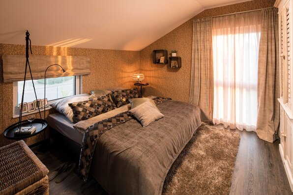 Schlafzimmer im Finger-Haus Musterhaus UNO 2.0 in Leipzig mit Doppelbett und gemusterter Bettwäsche, Dachschräge, großem Fenster mit Vorhängen, Teppich, Holzboden, Hängelampe und kleinen Wandregalen.