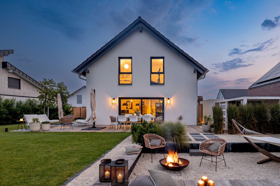 Abendstimmung im Garten eines modernen FingerHaus-Hauses mit gemütlicher Feuerstelle, stilvollen Sitzmöbeln, Hängematte und stimmungsvoller Beleuchtung.