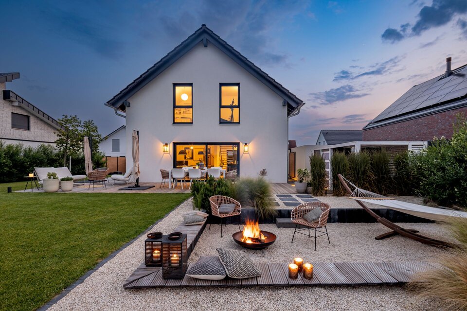 Abendstimmung im Garten eines modernen FingerHaus-Hauses mit gemütlicher Feuerstelle, stilvollen Sitzmöbeln, Hängematte und stimmungsvoller Beleuchtung.