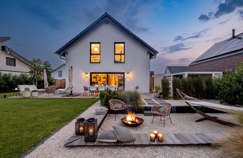 Abendstimmung im Garten eines modernen FingerHaus-Hauses mit gemütlicher Feuerstelle, stilvollen Sitzmöbeln, Hängematte und stimmungsvoller Beleuchtung.