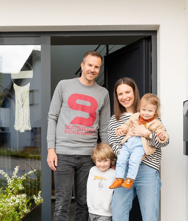 Eine Familie mit zwei kleinen Kindern steht fröhlich vor dem Eingang ihres modernen FingerHaus-Hauses. Die Eltern lächeln, während die Mutter das jüngere Kind auf dem Arm hält.