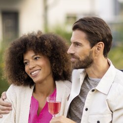 Ein Paar sitzt im Garten an einem gedeckten Tisch und genießt den Abend bei einem Glas Rosé. Beide wirken entspannt und lächeln, umgeben von sommerlicher Atmosphäre und grünem Hintergrund.