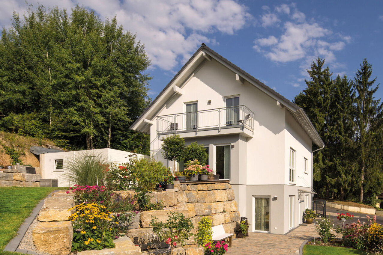 Weißes Einfamilienhaus mit Satteldach, Balkon und Untergeschoss an einem begrünten Hang mit Natursteinmauern.