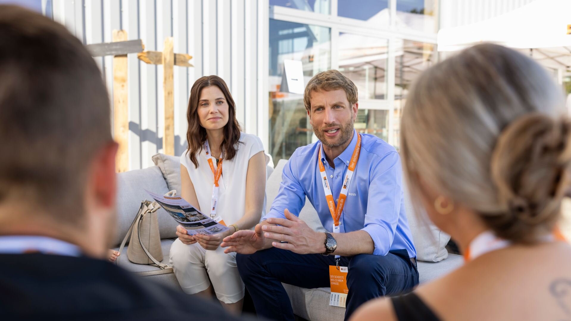 Zwei Personen mit orangen Namensschildern sitzen auf einem Sofa und sprechen mit zwei weiteren Personen, die mit dem Rücken zur Kamera zu sehen sind