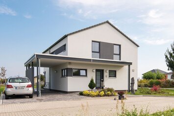Außenansicht des Finger-Haus Musterhauses MEDLEY 300 B in Berlin/Werder: zweigeschossiges Haus mit weißer Fassade und grauen Akzentflächen, Satteldach und Carport; davor gepflasterte Einfahrt mit geparktem Auto, Beetbepflanzung, Rasen und Bäume unter blauem Himmel.
