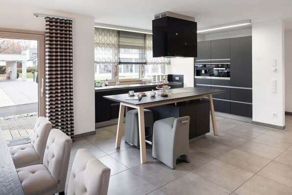 Moderne Küche im Musterhaus NEO 312 in Bad Vilbel mit dunklen Fronten, Kücheninsel mit integriertem Esstisch und stilvollen Sitzgelegenheiten.
