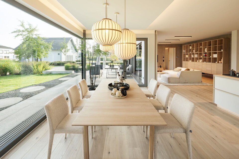 Offener Ess- und Wohnbereich im FingerHaus Musterhaus NIVO in Frankenberg mit Holztisch, gepolsterten Stühlen, Pendelleuchten und großer Fensterfront zum Garten.