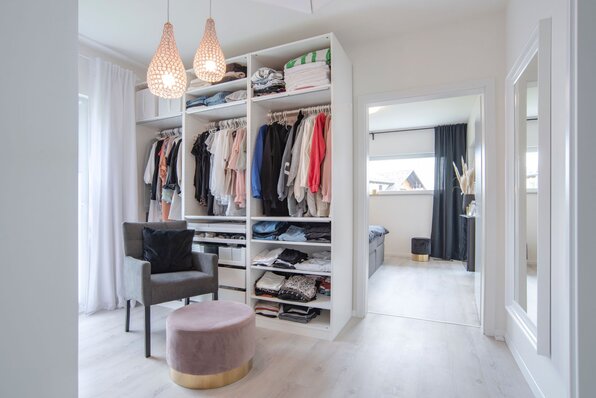Helle Ankleide in einem FingerHaus NIVO 130 mit offenem Kleiderschrank, Sessel, Hocker und Blick ins angrenzende Schlafzimmer.