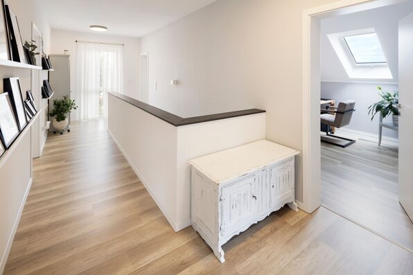 Heller Flur im Obergeschoss eines FingerHaus SENTO mit Holzfußboden, weißen Wänden und dekorativer Kommode; seitlich führt eine Tür in ein Zimmer mit Dachfenster.