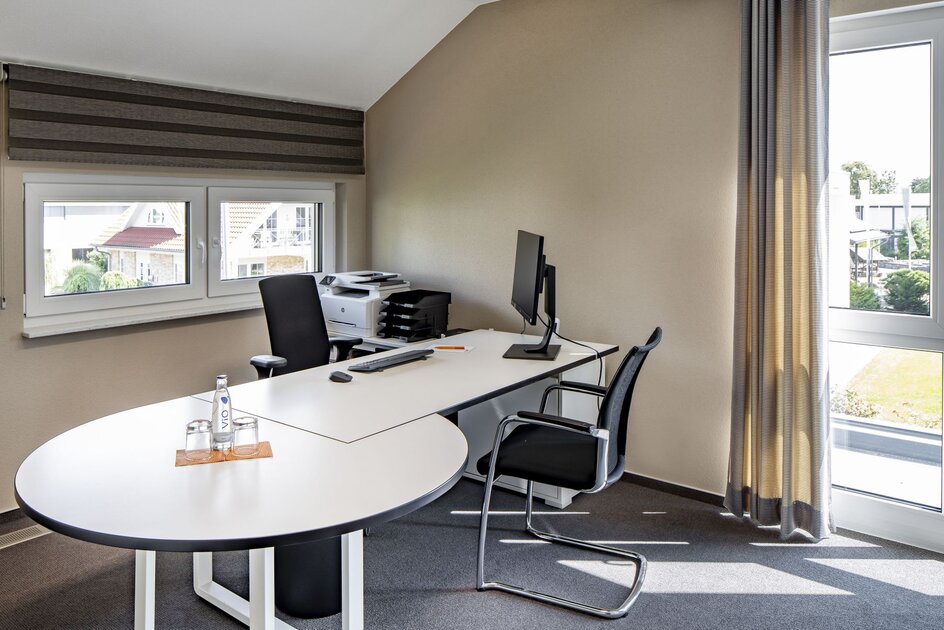 Modern eingerichtetes Büro mit Schreibtisch, Computer, Drucker und zwei Stühlen in einem hellen Raum mit großen Fenstern und grauen Vorhängen.