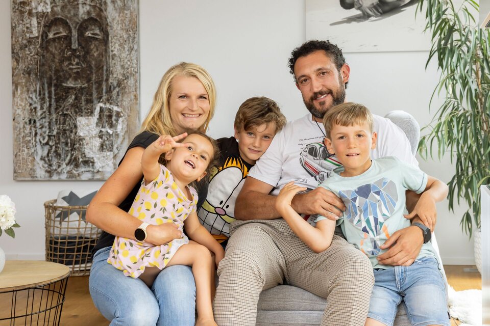 Eine fünfköpfige Familie mit zwei Erwachsenen und drei Kindern sitzt gemeinsam in einem Wohnraum und blickt in die Kamera.