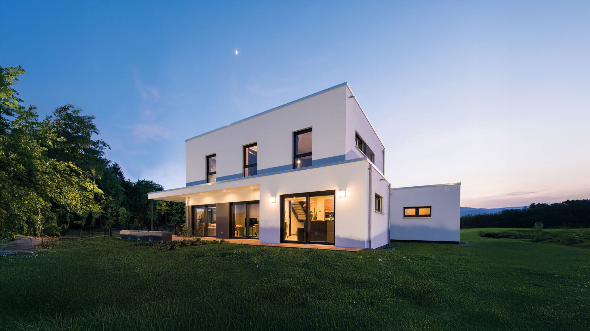Modernes, zweigeschossiges Flachdachhaus mit weißer Fassade und großen Fensterflächen, seitlichem Anbau (Einliegerwohnung), überdachter Terrasse und weitläufiger Rasenfläche, aufgenommen in der Abenddämmerung mit blauem Himmel.
